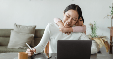 woman at computer with child's arms wrapped around her lovingly