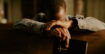 man with head down and crossed hands holding Bible